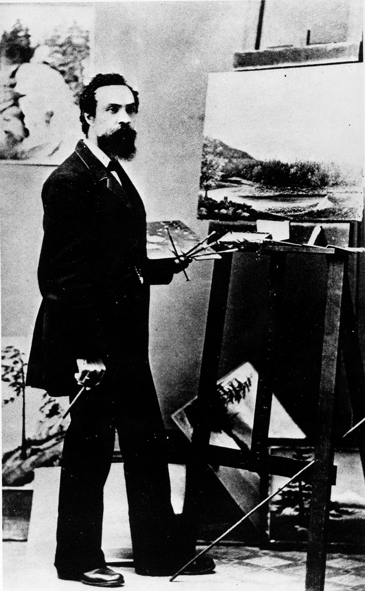 A black-and-white photo of a man in a suit, holding a palette and brushes in front of an easel.